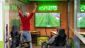 Un área de juegos con una pantalla grande mostrando un partido de fútbol, sillas gamer y una pared de ladrillo con un letrero de neón que dice "eSPORTS".