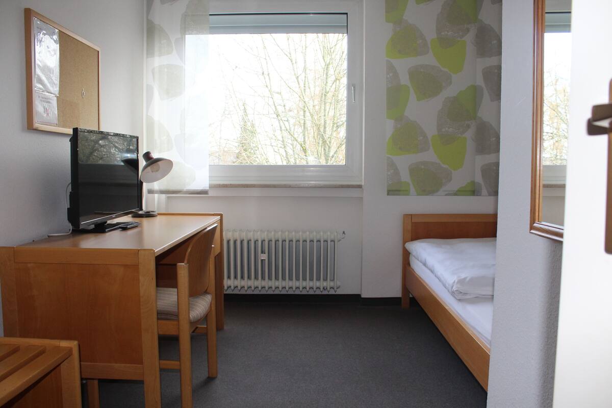 A small, tidy room with a wooden desk, a chair, a bed, a radiator, and a window with a view of trees.