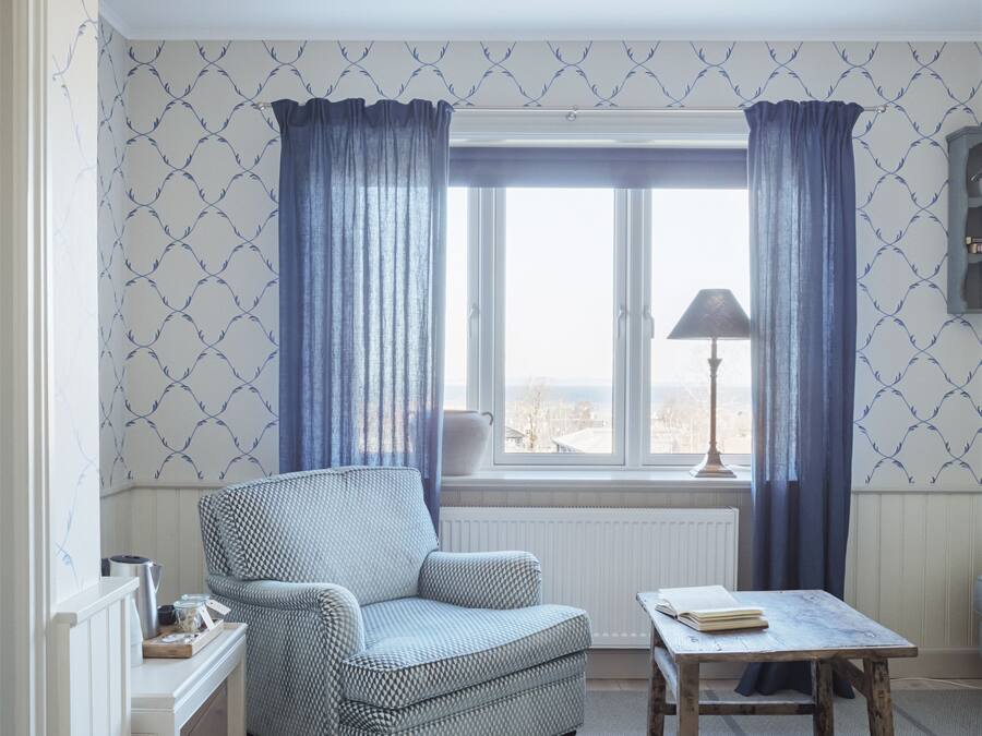 Una habitación con papel tapiz estampado, un sillón azul, una mesita y vistas a través de una ventana.