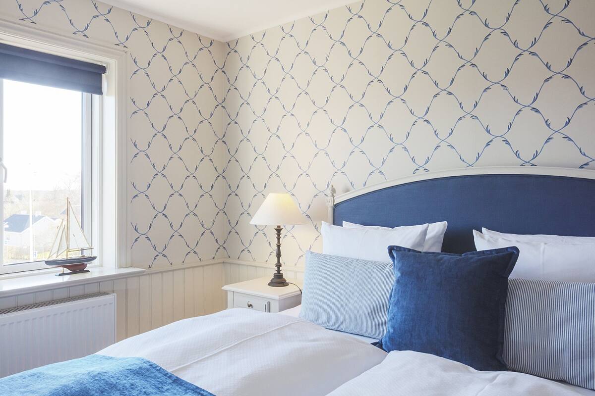 Un lit bien fait, avec une literie blanche et bleue, une table de chevet avec une lampe, et une fenêtre donnant sur des immeubles.