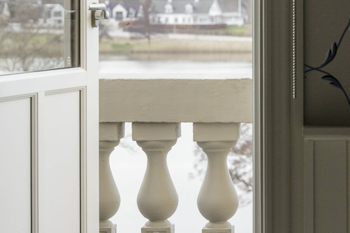 Une vue depuis une fenêtre ornée d’une balustrade, donnant sur une étendue d’eau et sur des maisons situées au loin.