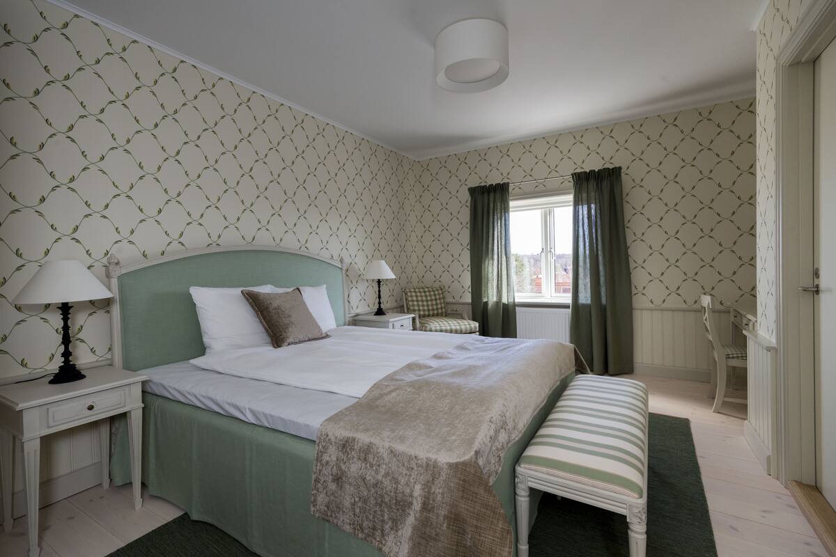 Une chambre bien rangée, avec un lit, des tables de chevet, un banc et une fenêtre avec des rideaux.