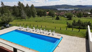 A swimming pool with lounge chairs and tables, surrounded by a grassy area with trees and a view of a village and mountains in the distance. - Siljansnäs Hotell (Siljansnas)