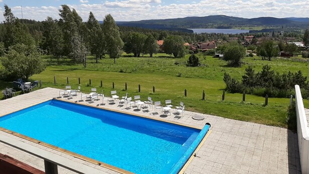 Una piscina con sillones y mesas, rodeada de césped, árboles y vistas a un pueblo y montañas a lo lejos.