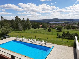 Una piscina con sillones y mesas, rodeada de césped, árboles y vistas a un pueblo y montañas a lo lejos.