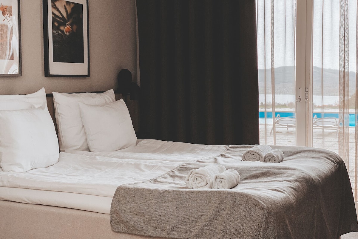 A hotel room with a bed, white pillows, a dark curtain, and a view of a pool through a large window.