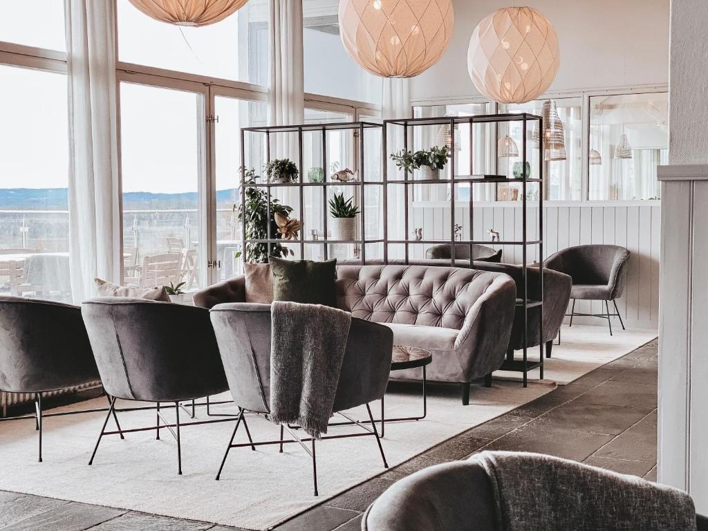 A modern interior with a tufted sofa, black chairs, a plant-filled shelving unit, and large windows offering a city view.