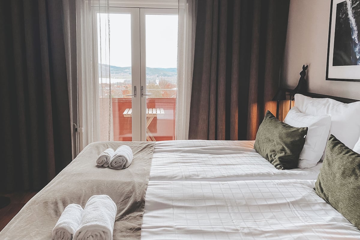 A hotel room with a bed, two rolled towels, a view through a glass door, and a framed picture on the wall.