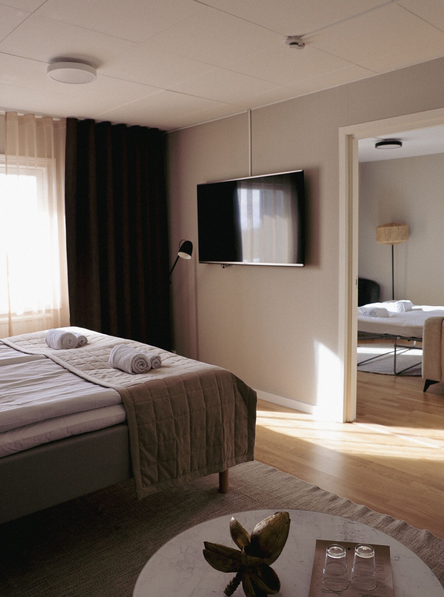 A hotel room with a bed, a flat-screen TV, a bedside lamp, and a small table with glasses.