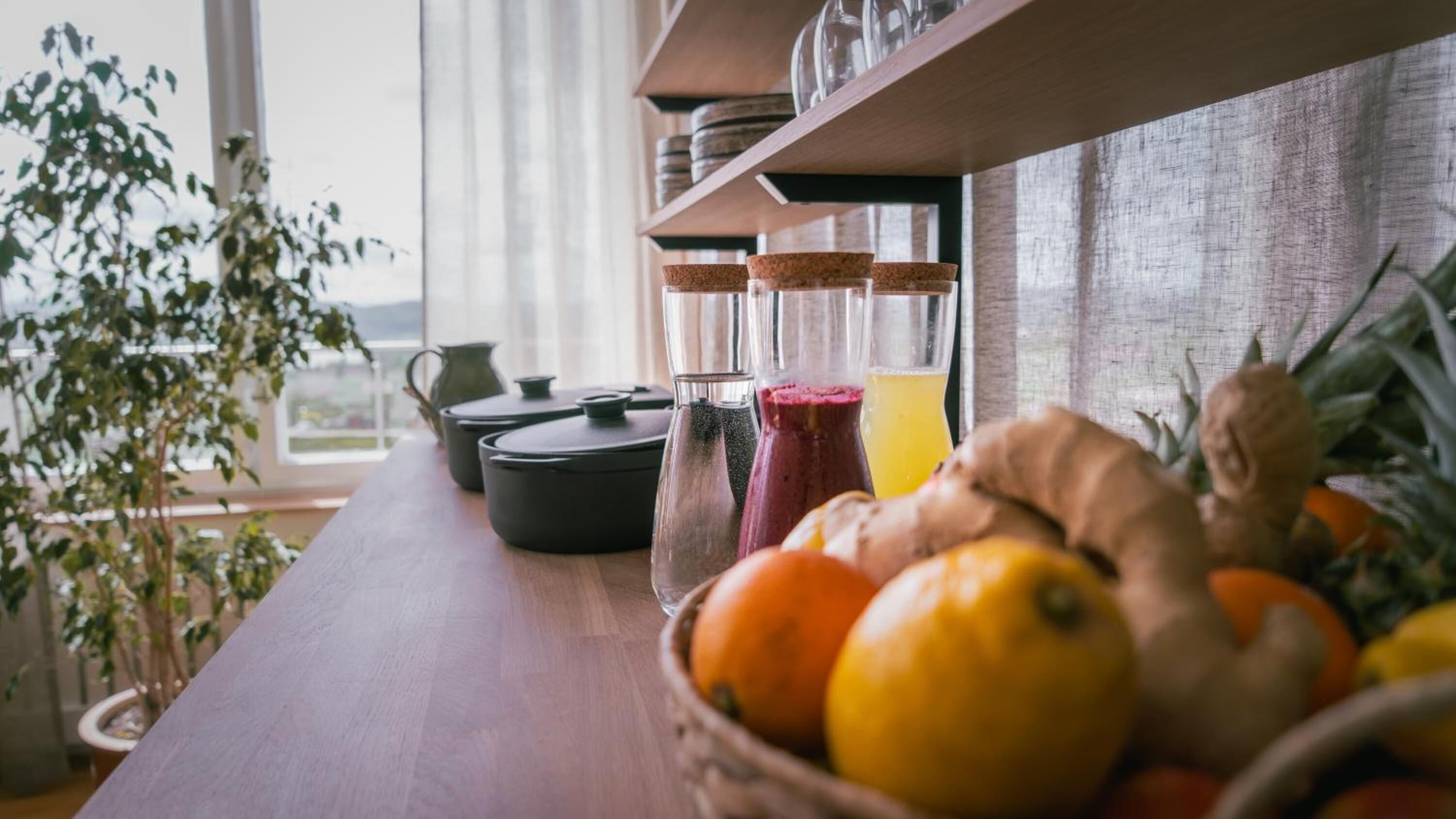 Una encimera de cocina con un bol de frutas, una jarras y un vaso de jugo de naranja.