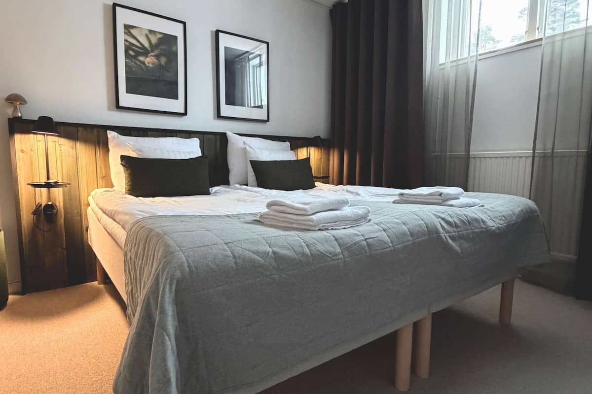A neatly made bed with white linens and folded towels, a wooden headboard, and two framed pictures on the wall.