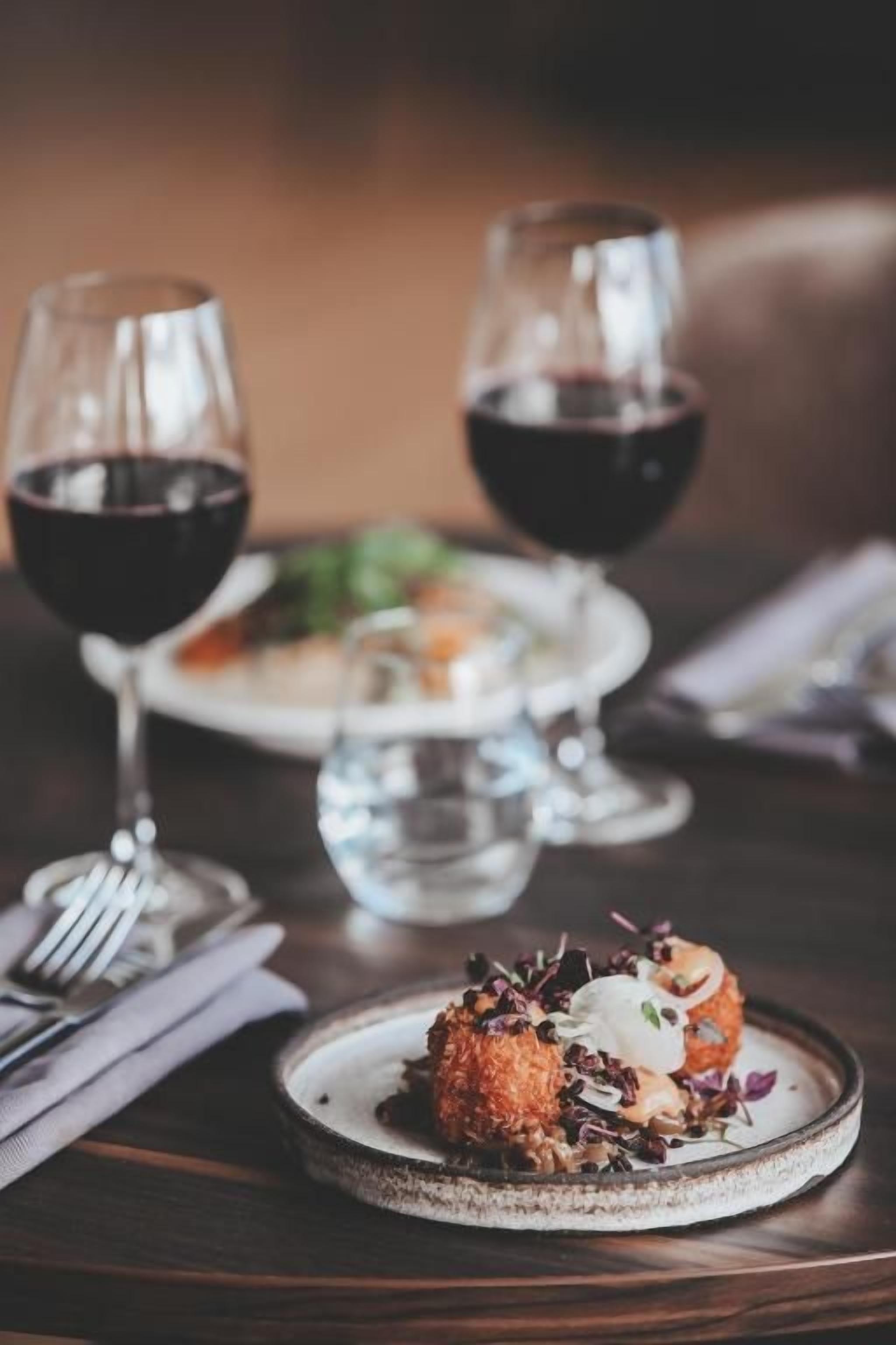 A dining table with a plate of food, two glasses of red wine, and a fork on a napkin.