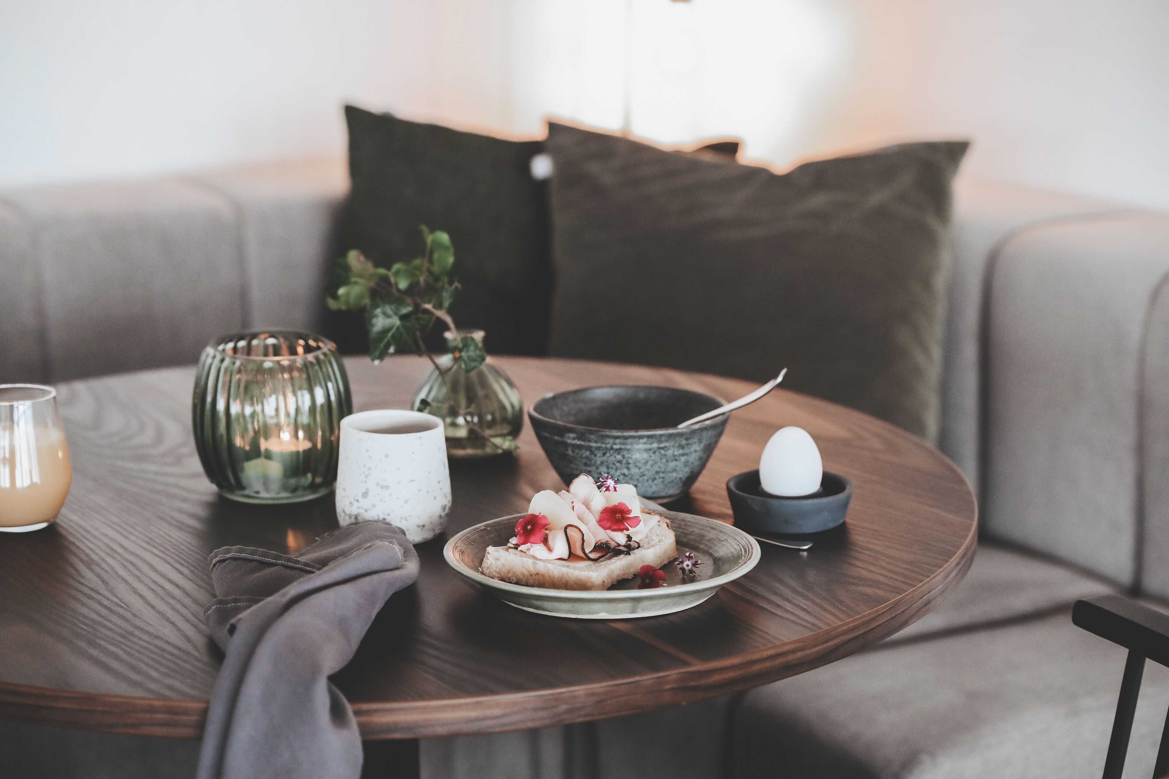 A round wooden table with a plate of toast topped with fruit and a boiled egg, a glass of orange juice, a candle, and a vase with a plant.