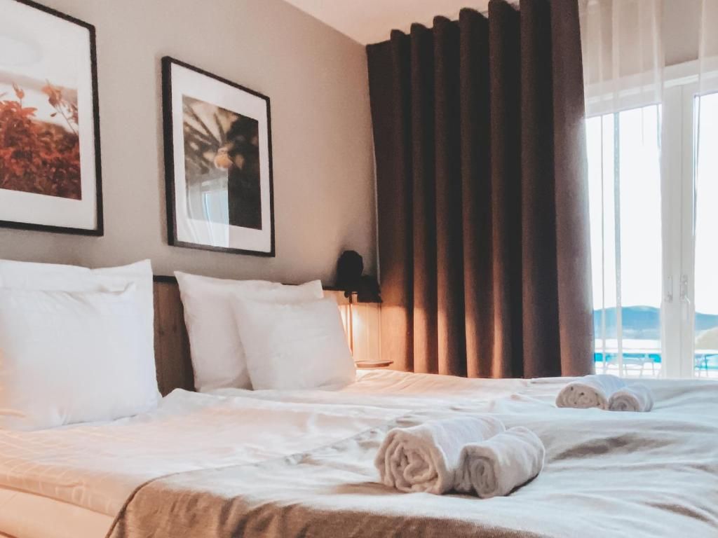 A hotel room with a bed, pillows, a nightstand, a lamp, and a window with curtains.