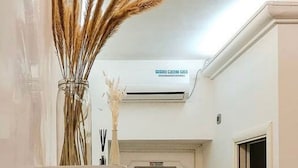 Un couloir étroit avec une porte blanche, un climatiseur fixé au mur et un vase contenant des plantes séchées.