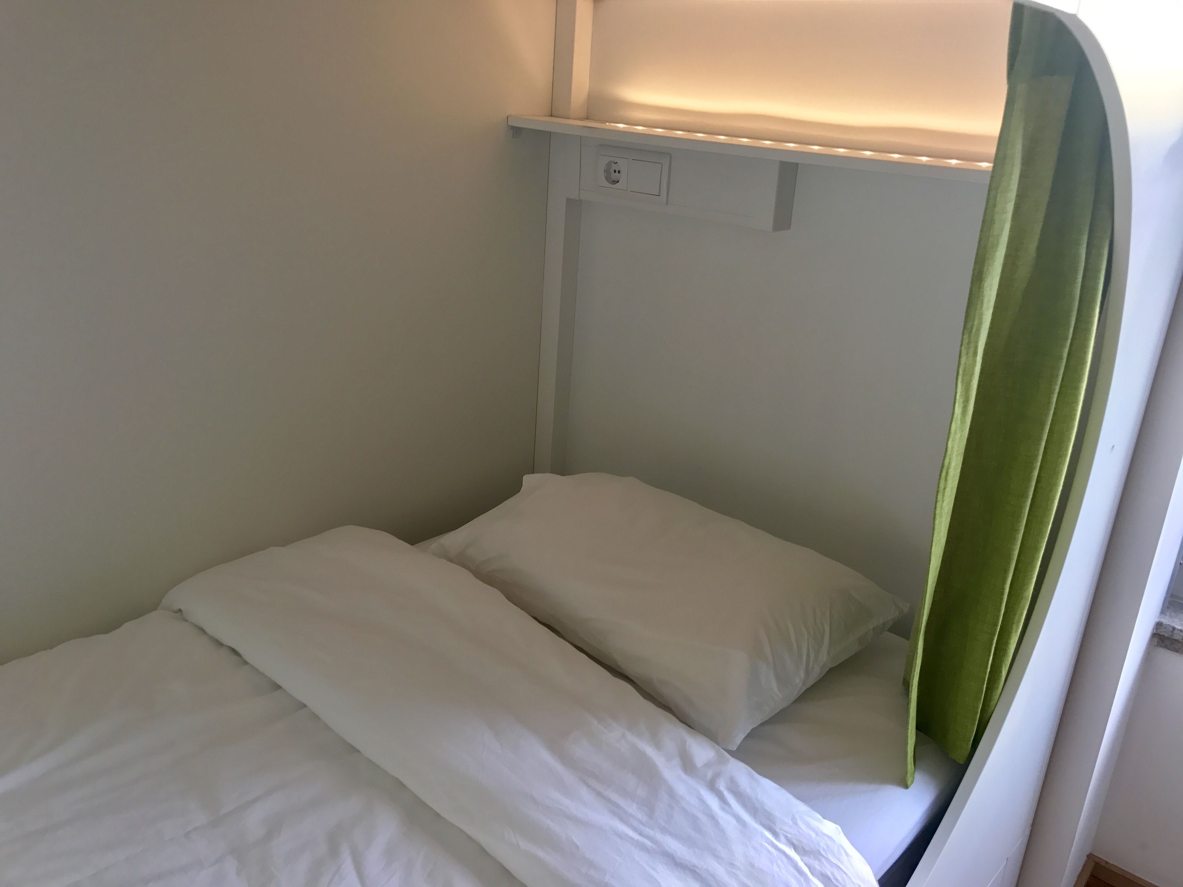 A small, single-bed room with a green curtain, white bedding, and a wall-mounted light fixture.