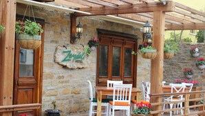 A stone building with a wooden pergola, a wooden table with chairs, and hanging flower baskets. - Zeydali Hotel (Gökçeada)