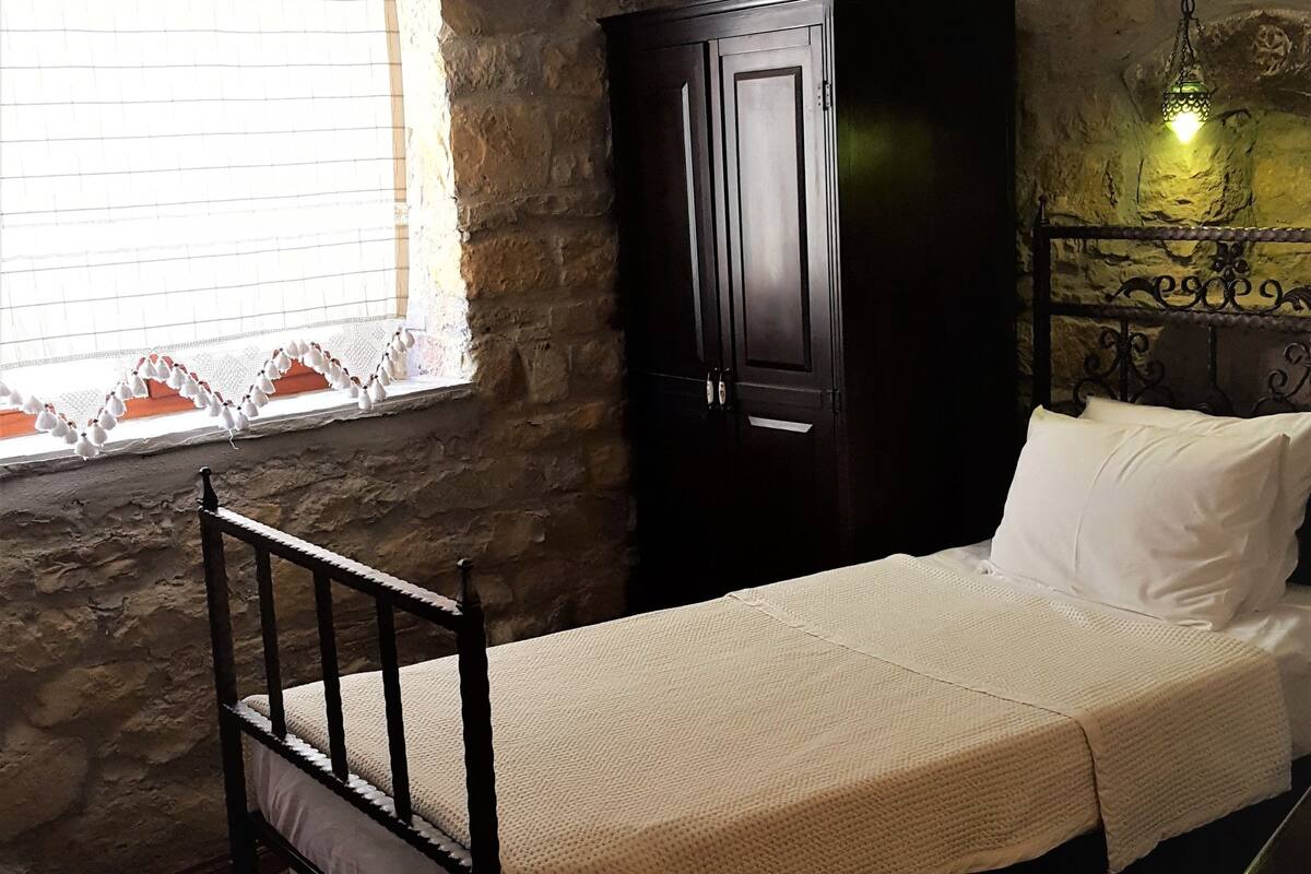 A bedroom with a wooden bed, a stone wall, a window with a curtain, and a wall-mounted lamp.
