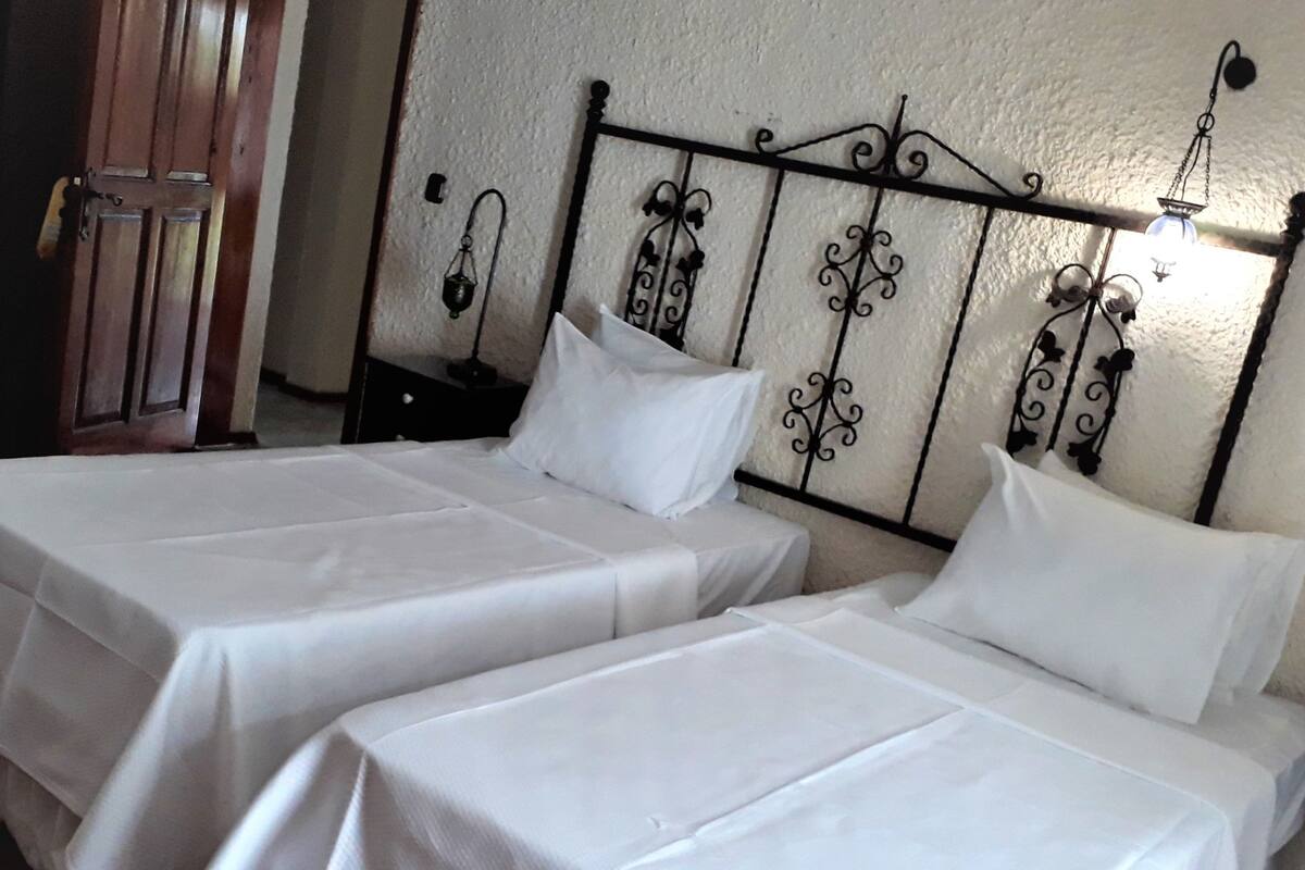 A room with two single beds, each with a white bedspread and pillow, against a textured wall with a decorative metal headboard.