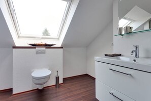 Un baño moderno con tragaluz, inodoro blanco, repisa de vidrio con libros y tocador blanco con espejo.
