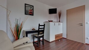 A room with a wooden floor, a white desk with a chair, a television mounted on the wall, and a cabinet with a vase of dried plants. - Pension Haselmann (Münster)