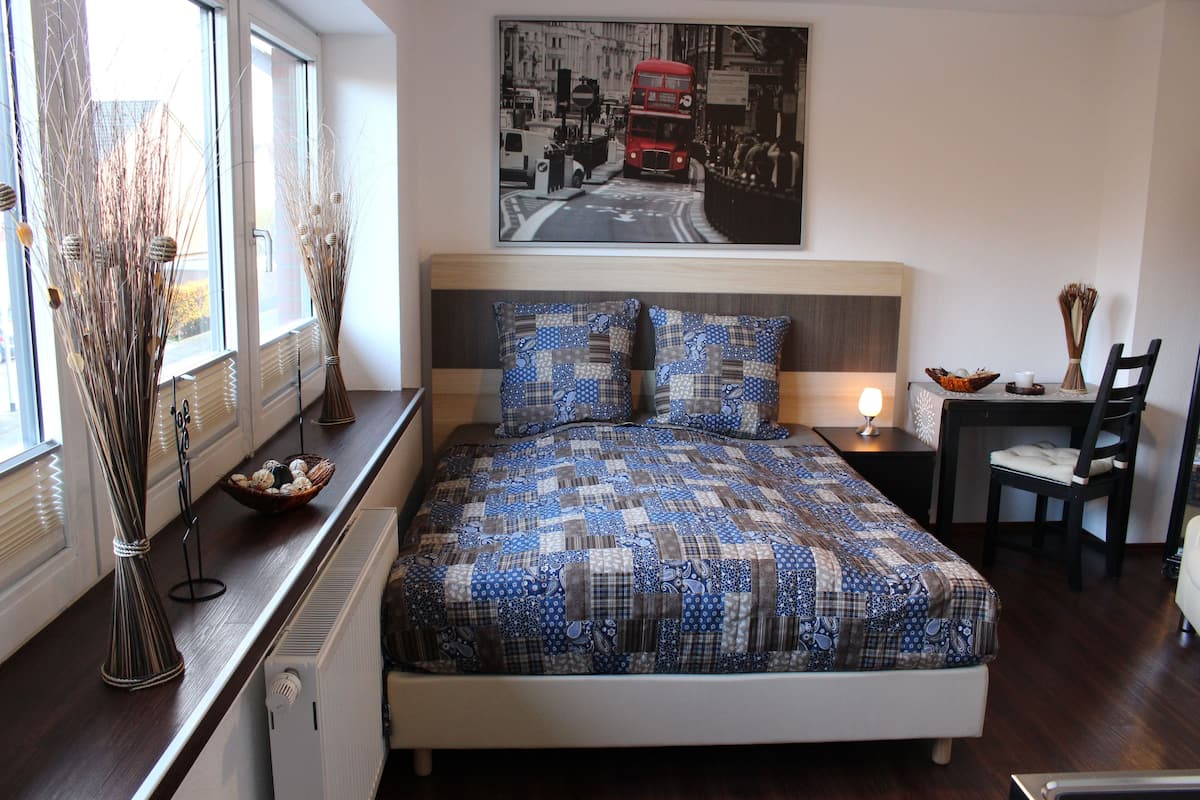 Quarto com cama, janela com vista para uma rua da cidade, mesa de cabeceira com abajur e uma cadeira.