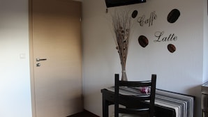 A room with a wooden door, a table with a striped tablecloth, a chair, a vase with dried plants, and a wall with the words "Caffe Latte" and coffee beans. - Pension Haselmann (Münster)