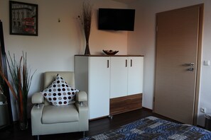 Una habitación con un sillón color crema, un armario blanco, una puerta de madera y un televisor de pantalla plana en la pared.