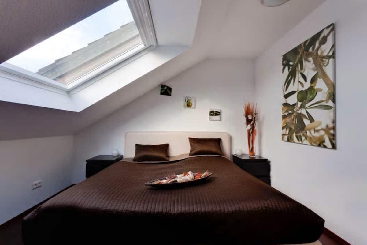 Quarto com claraboia, uma cama grande, mesa de cabeceira, obra de arte na parede e uma pequena planta.
