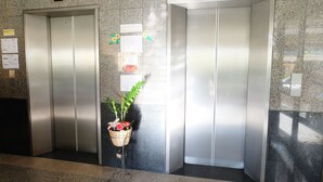 Two elevators in a lobby with a digital display above each, a plant in a basket, and informational signs on the wall. - Grand Living Place (Bangkok)