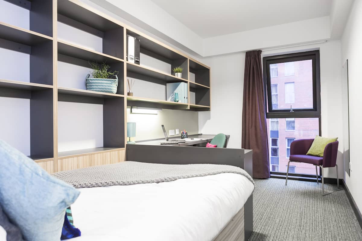 A modern hotel room with a large bed, built-in shelving, a desk with a laptop, and a view of a building through a window.