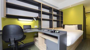 A hotel room with a bed, a desk with a laptop, a chair, and a shelf unit.