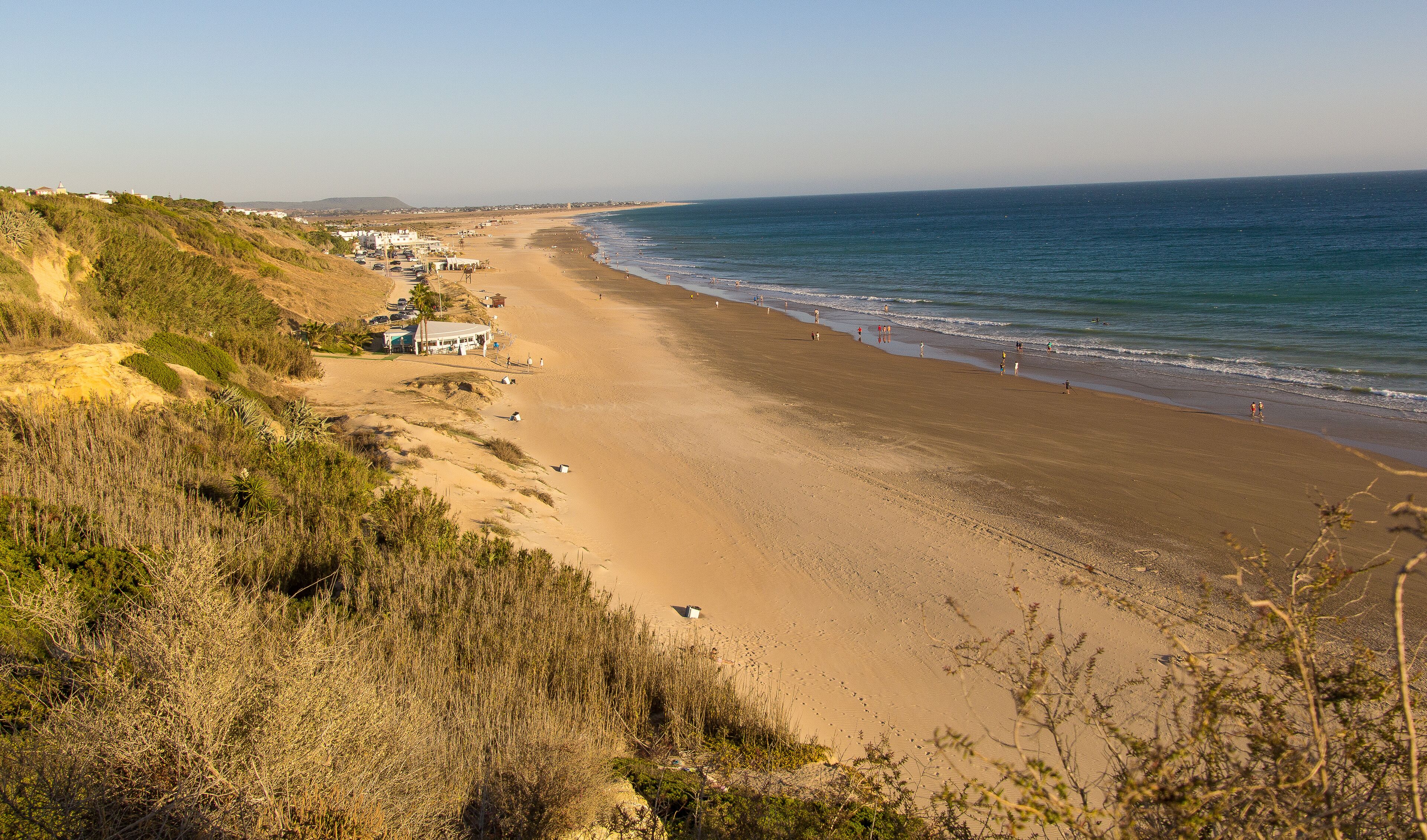 A coastal beach with a sandy shore, a few buildings, and a clear sky.