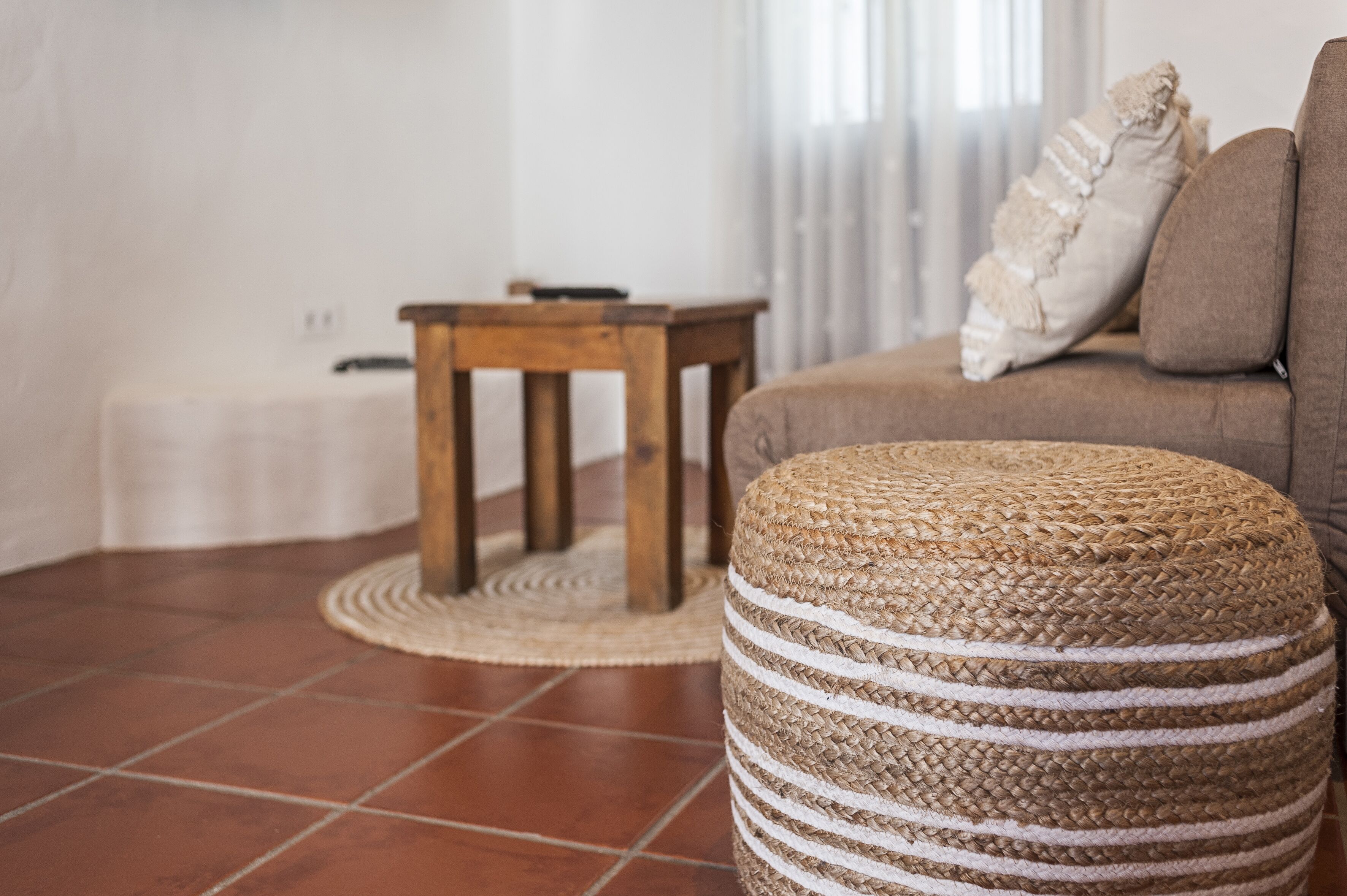 Un salon avec un canapé marron, un pouf rond en fibres tressées, une petite table en bois et un tapis blanc.