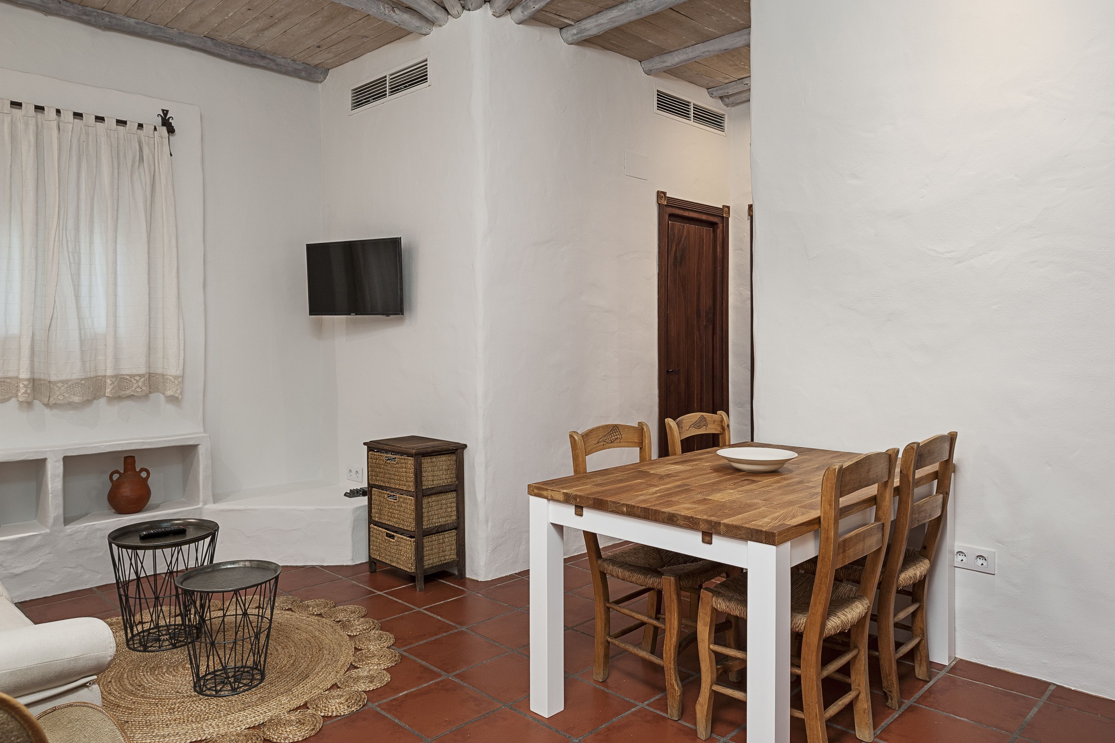 Un comedor con una mesa y sillas de madera, un televisor montado en la pared y una ventana con cortinas.