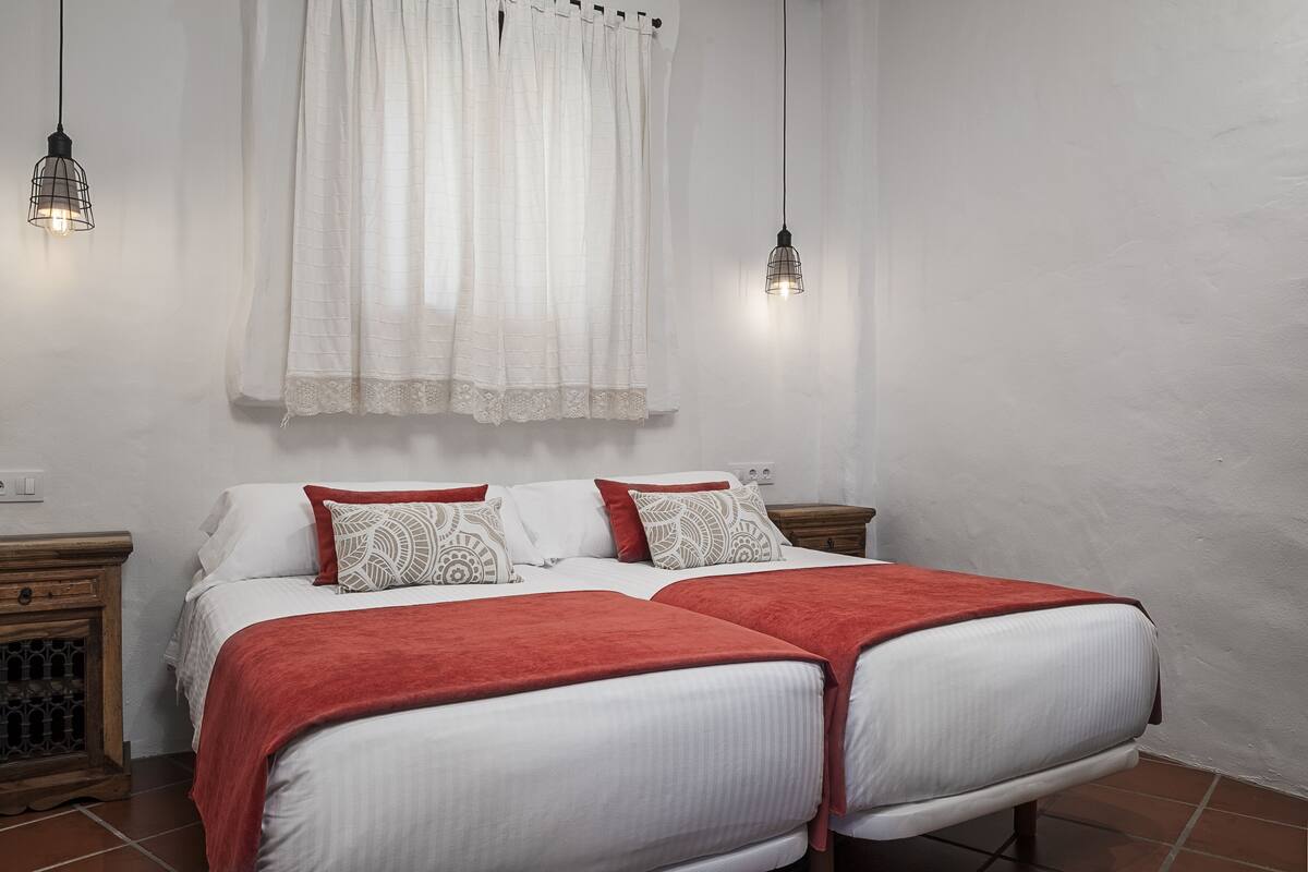 Una cama doble con ropa de cama blanca y roja, dos mesitas de noche y una ventana con cortinas blancas.
