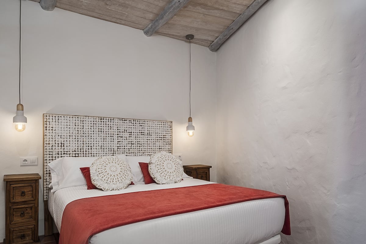 Un dormitorio con cabecera de madera, colcha roja y dos mesitas de noche con lámparas.