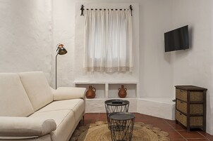 A living room with a sofa, a small table with a lamp, a TV mounted on the wall, and a decorative window with curtains.