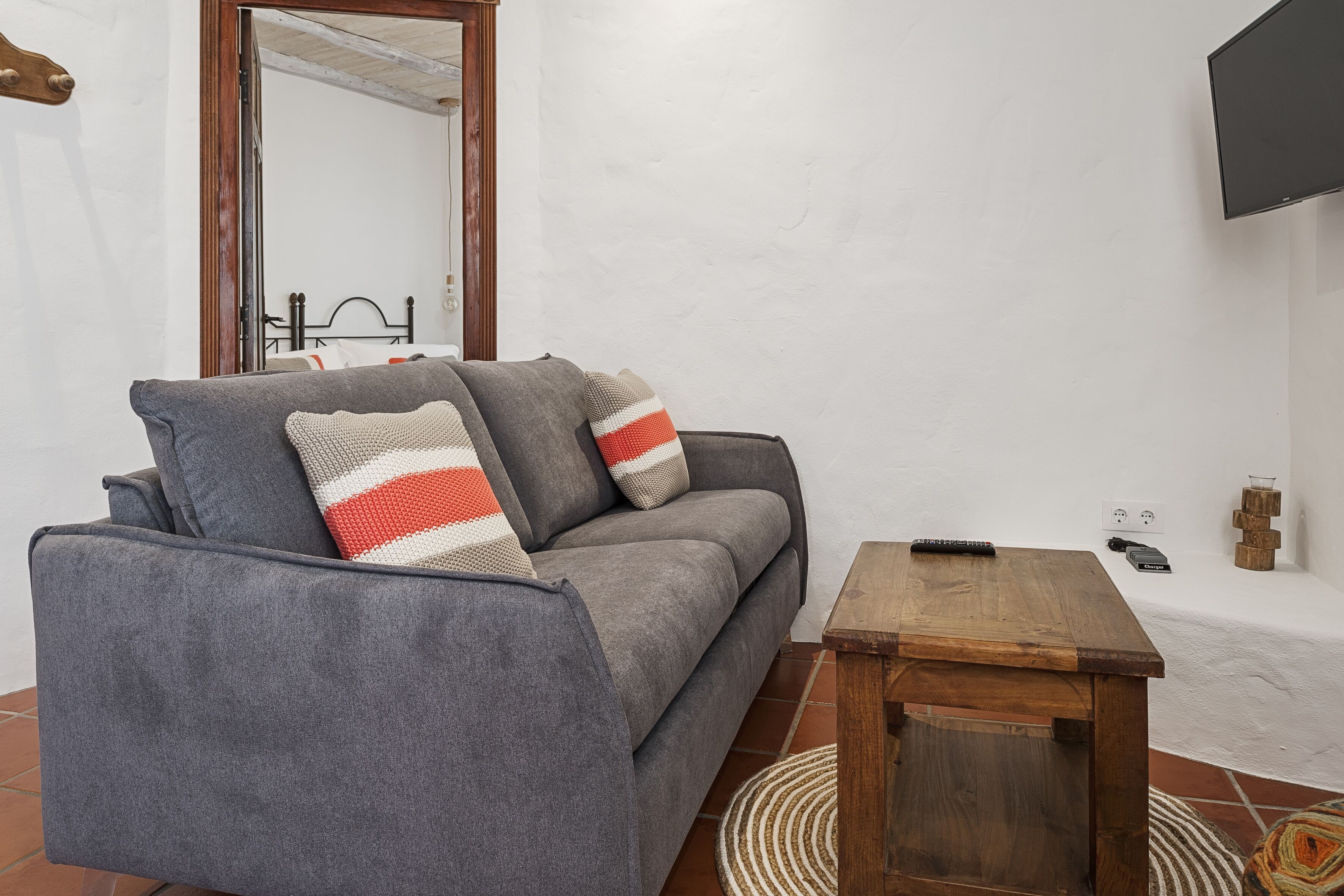 Un sofá gris con almohadones a rayas, una mesita de madera, un televisor montado en la pared y un piso de baldosas.