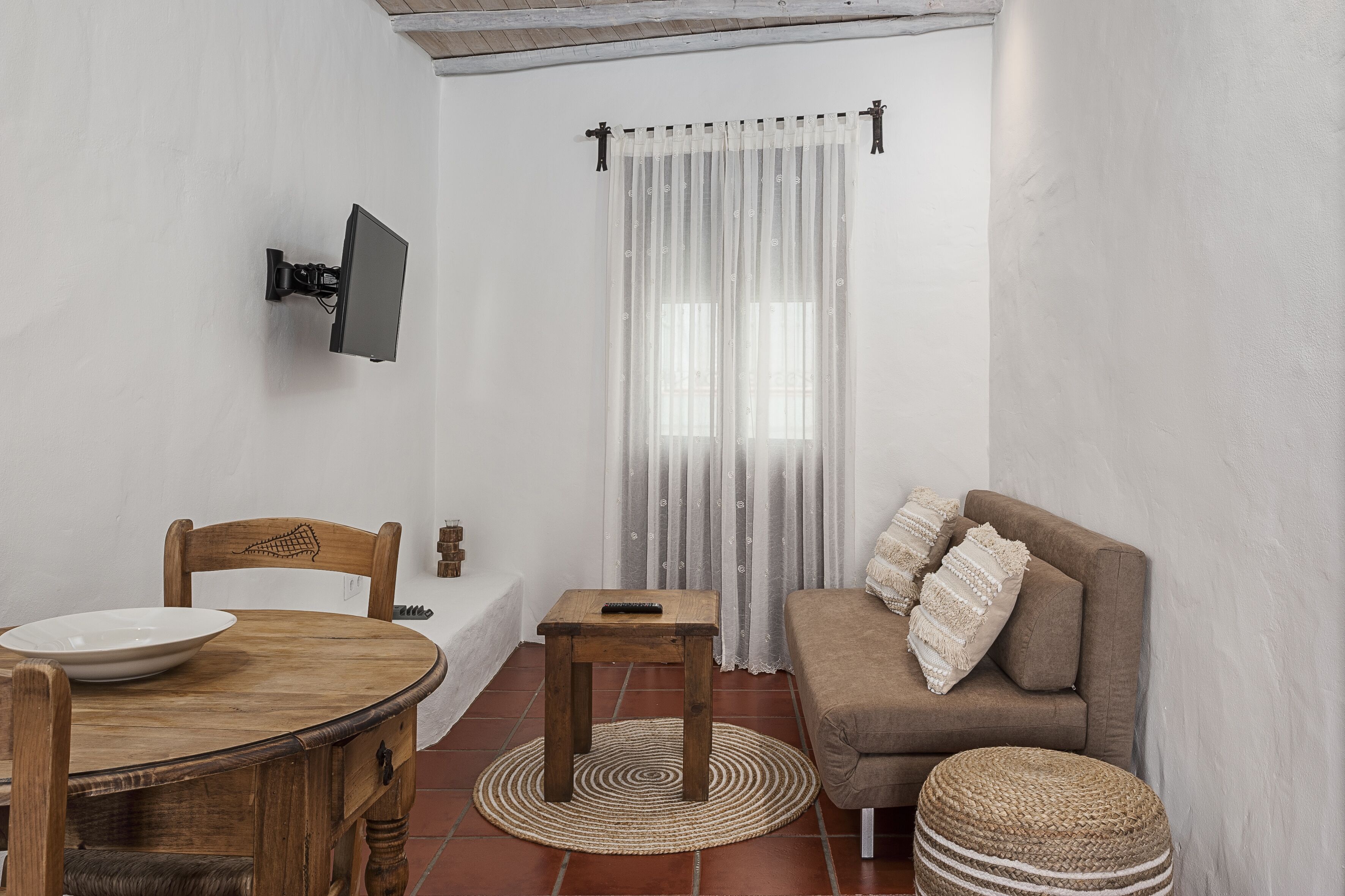 Una habitació amb taula de dinar de fusta, un sofà, un televisor fixat a la paret i una finestra amb cortines de tela fina.