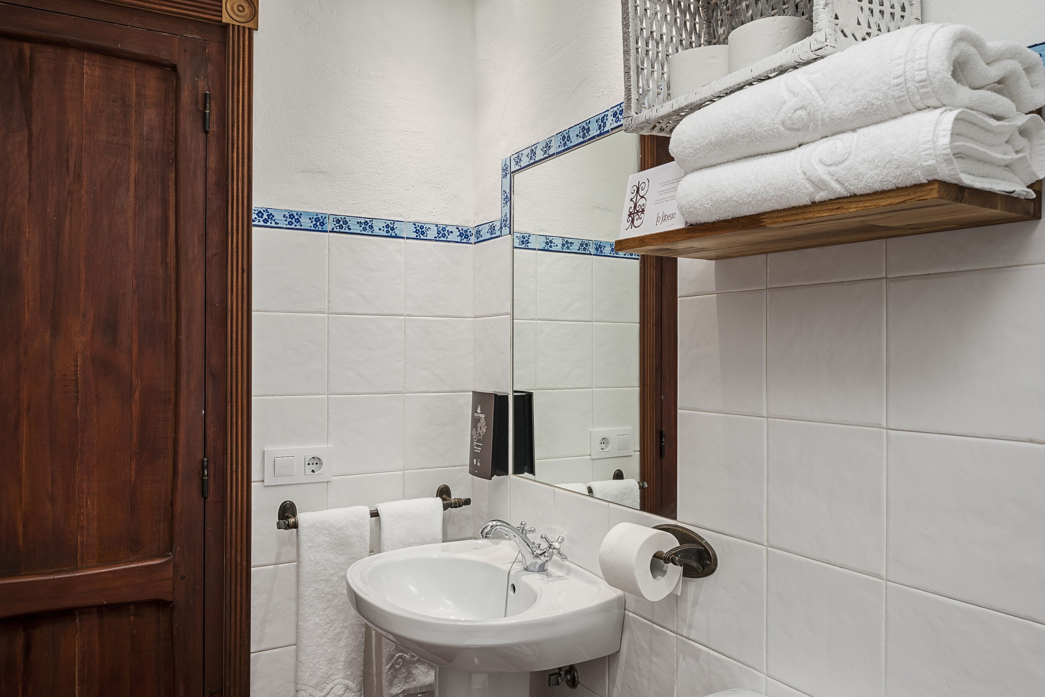 A bathroom with a wooden door, white tiles, a sink, a mirror, and a towel rack with folded towels.