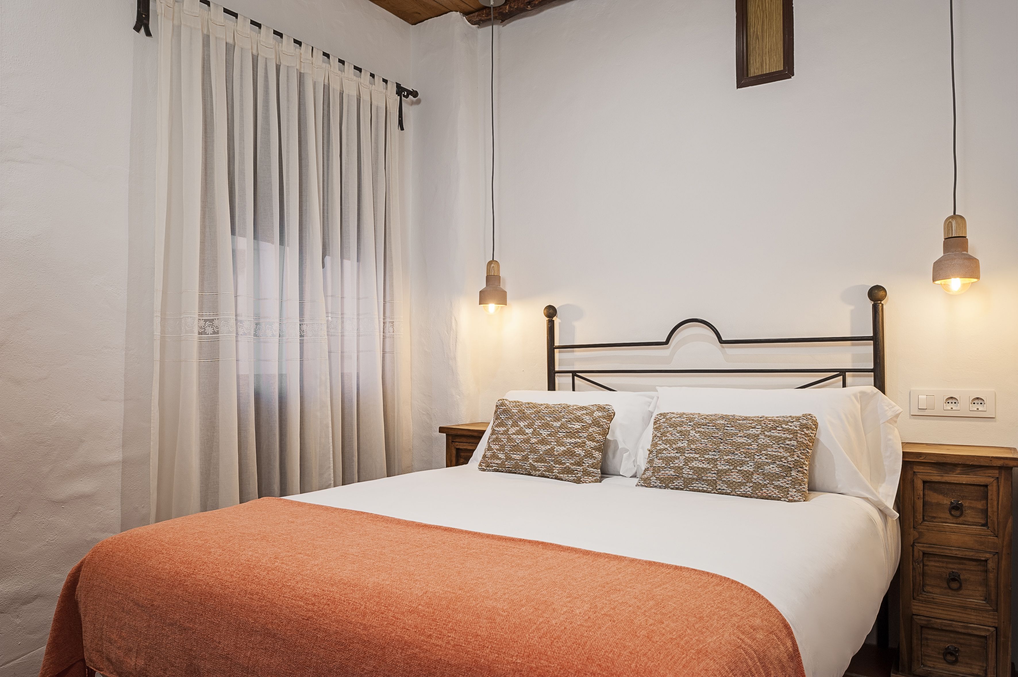 A neatly made bed with a white and orange blanket, two patterned pillows, and a wooden headboard. There are two bedside lamps and a small wooden nightstand with drawers.