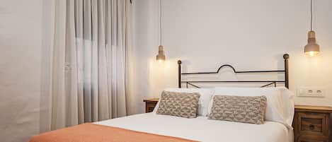 A neatly made bed with a white and orange blanket, two patterned pillows, and a wooden headboard. There are two bedside lamps and a small wooden nightstand with drawers.