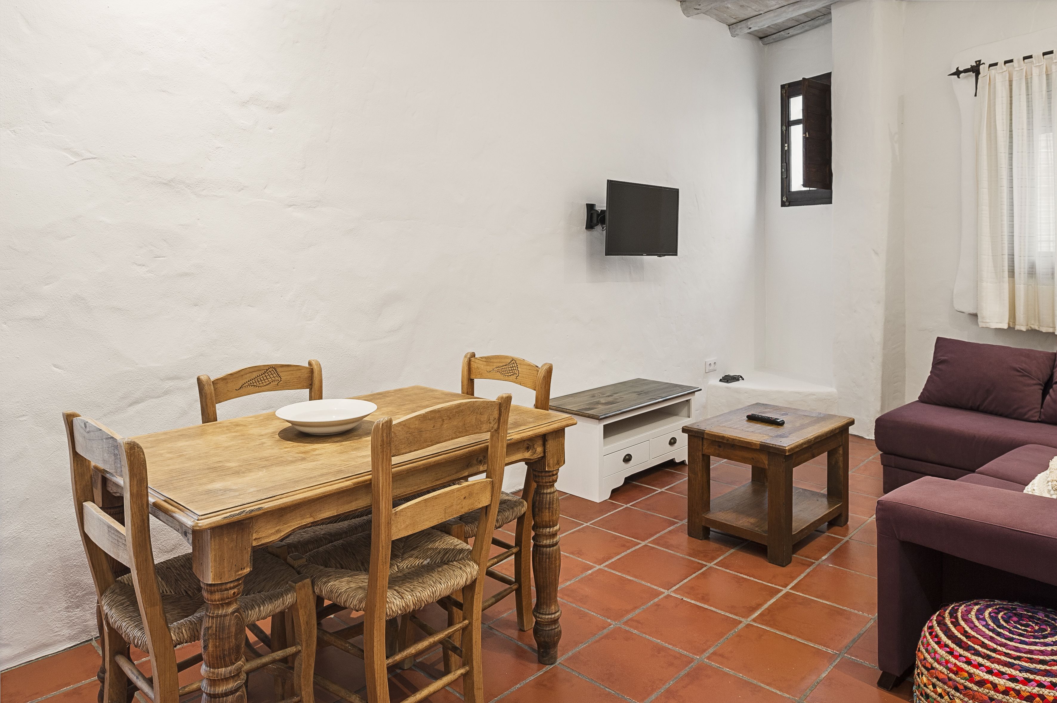 Un espace de vie compact comprenant une table à manger en bois, des chaises, une petite table d’appoint, un canapé et une fenêtre avec un rideau.