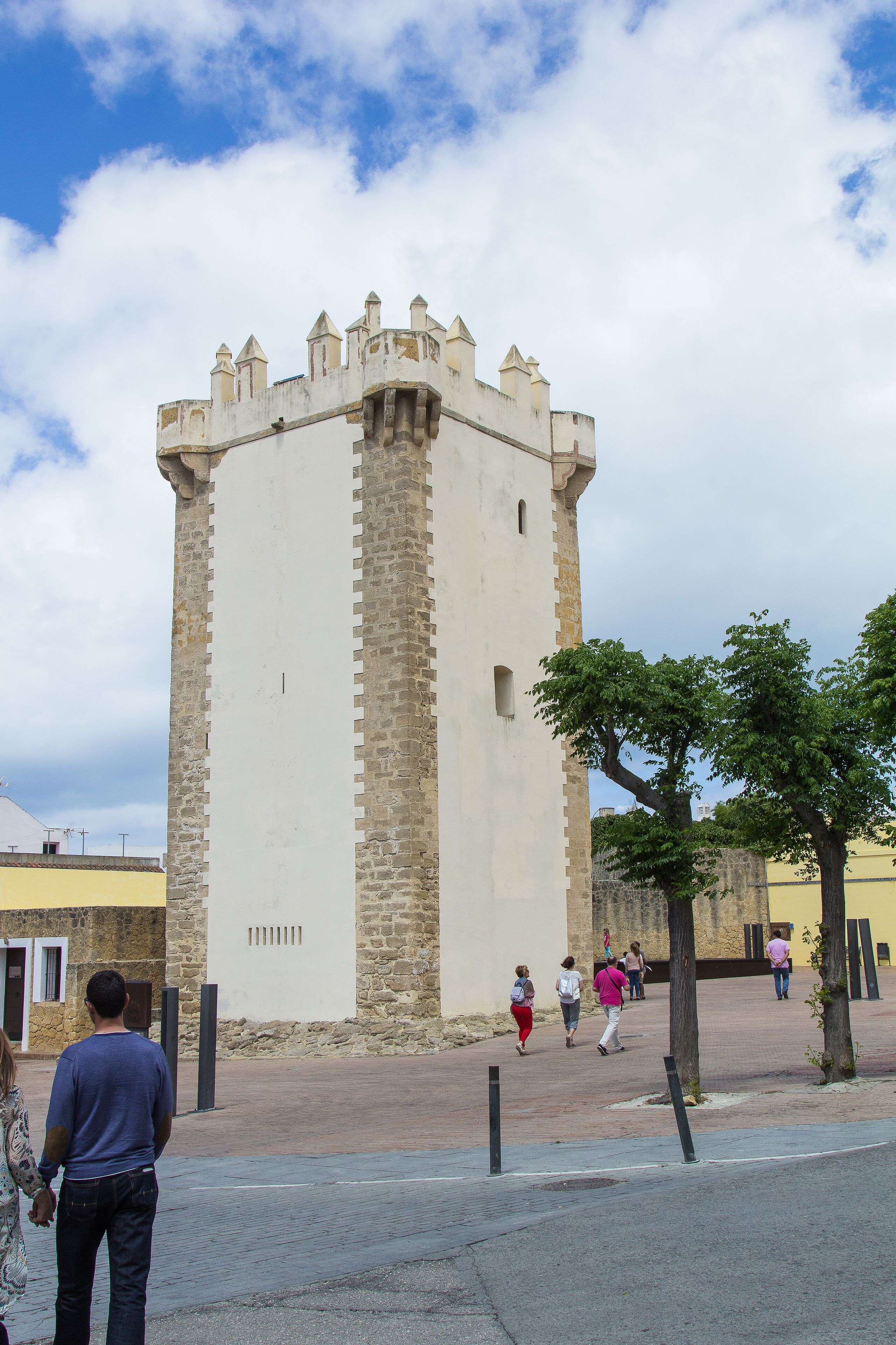 Una torre histórica de base cuadrada y almenas, rodeada por una zona pavimentada con árboles y algunas personas caminando.