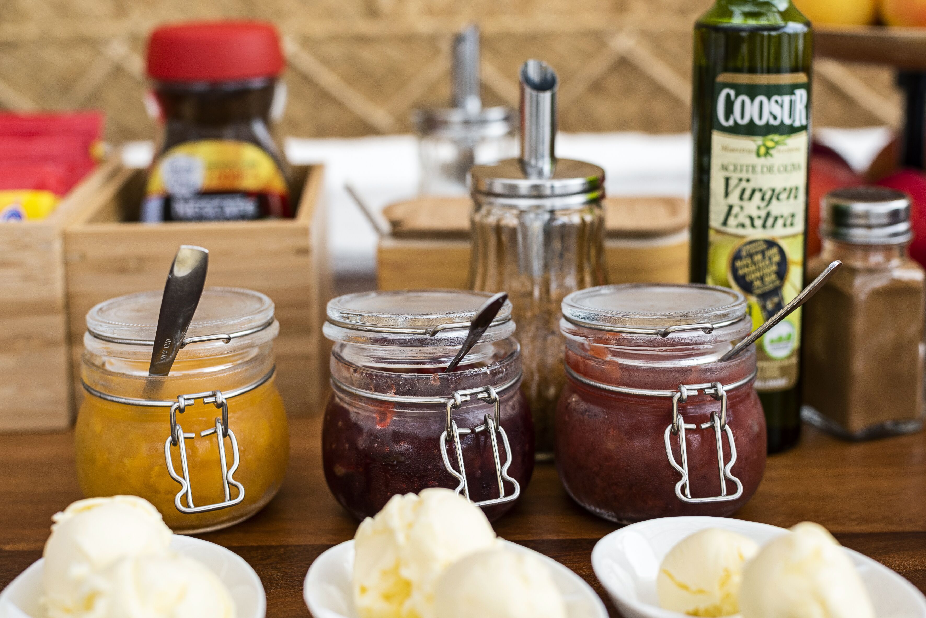 Three jars of preserves with a spoon in each, a bottle of Coosur, and a wooden crate in the background.