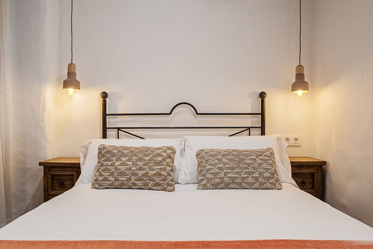 Una cama bien hecha con almohadones estampados, dos mesitas de noche y lámparas colgantes.