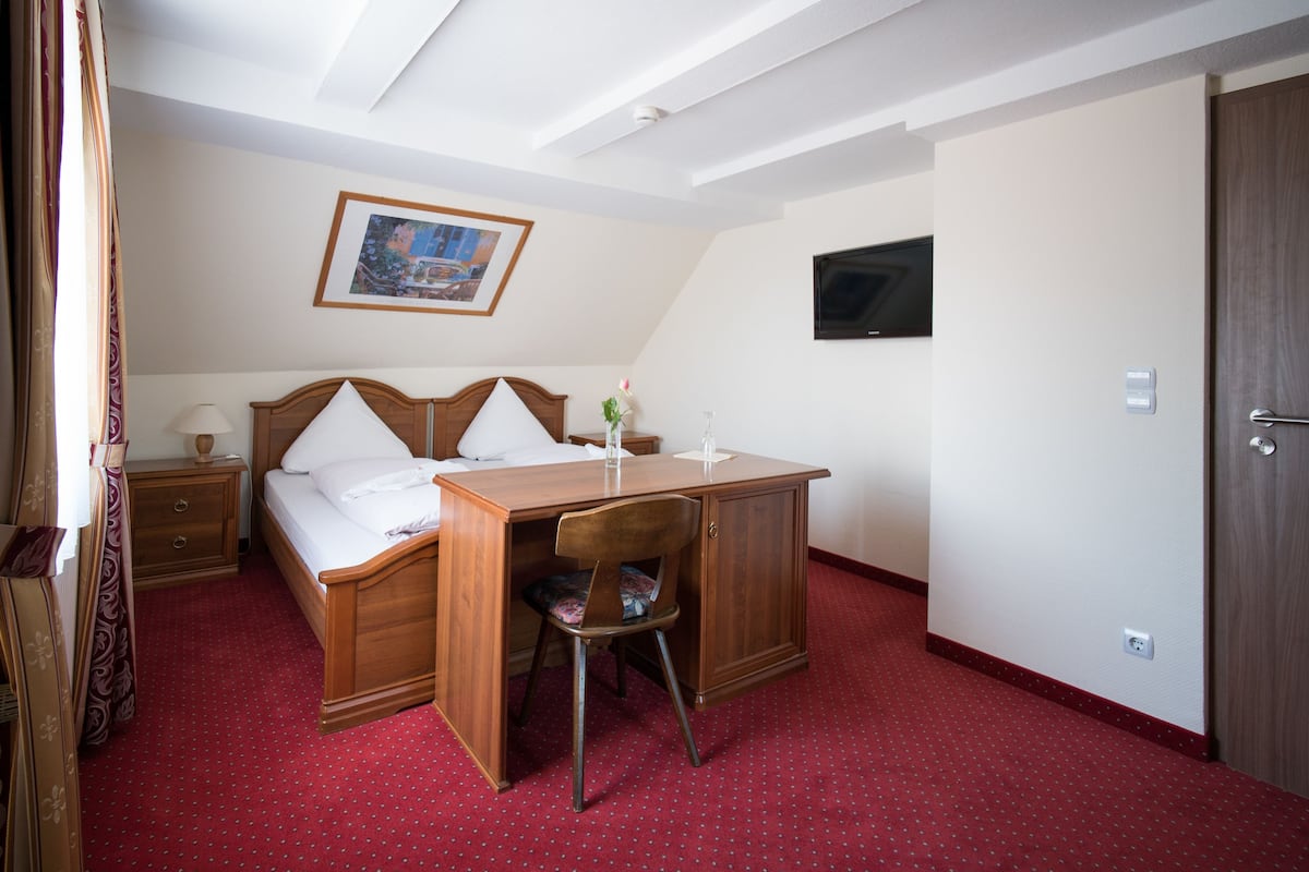 A hotel room with a bed, desk, chair, nightstand, lamp, and a painting on the wall.