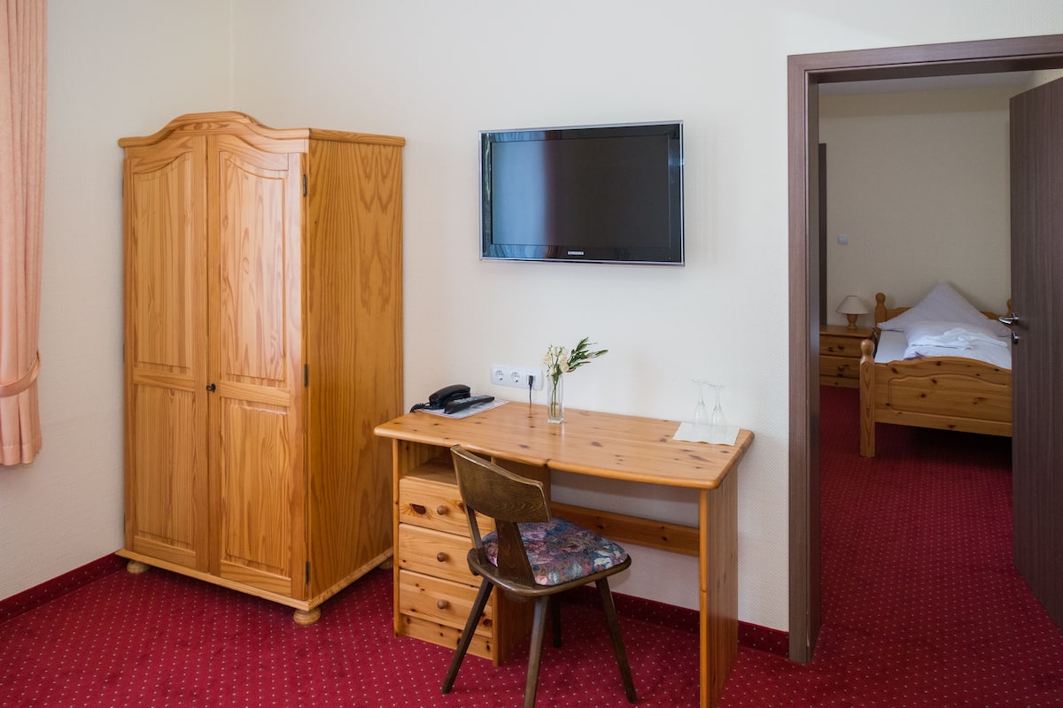 A room with a wooden desk, a chair, a wardrobe, a television, and a bed.