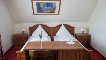 Uma cama de casal com roupa de cama branca, duas mesas de cabeceira com abajures, uma escrivaninha de madeira, uma cadeira e um quadro na parede.
